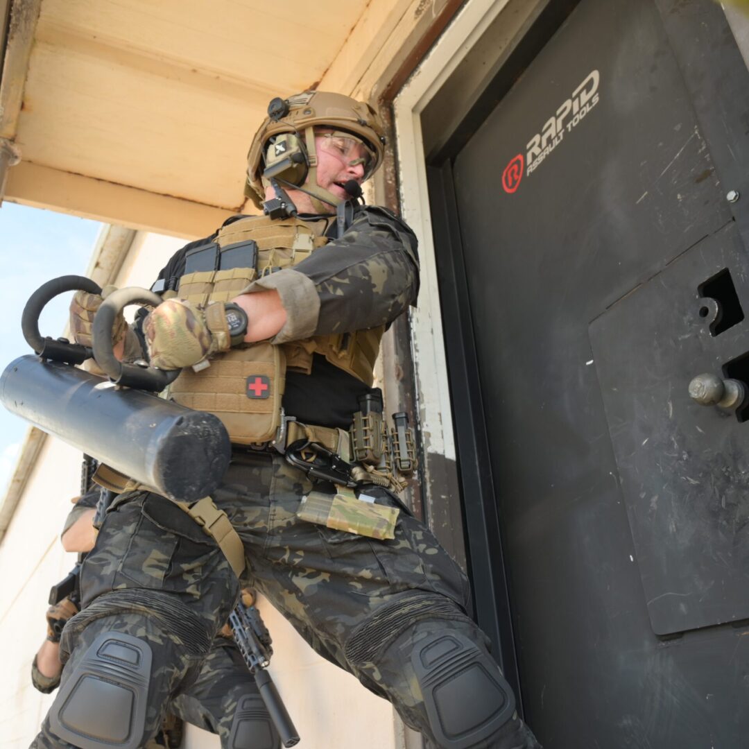 A soldier in camouflage holding a gun and standing next to a door.