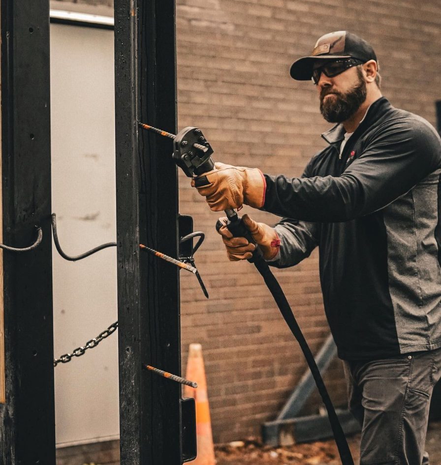 A man holding an electric drill near a metal structure.